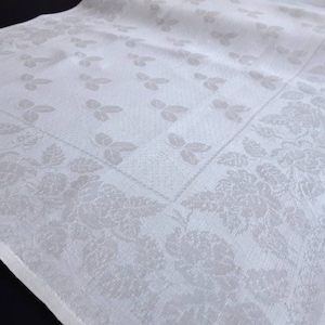 May include: White linen tablecloth with a damask pattern of leaves and flowers. The tablecloth has a border of a repeating floral design.