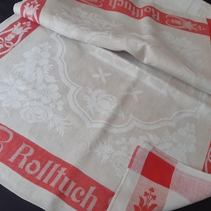 Pode incluir: Toalha de mesa de linho creme com um padrão floral e borda vermelha. A borda apresenta a palavra "Rolltuch" e elementos decorativos. Uma pequena guardanapo xadrez vermelho e branco é visível.