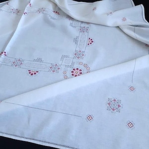 Pode incluir: Toalha de mesa de linho branco com desenhos florais e geométricos bordados em tons de rosa e cinza.