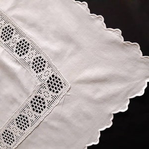 Könnte beinhalten: Eine weiße Baumwolltischdecke mit einem gewellten Rand und einer zarten Spitzenborte. Die Spitze zeigt ein sich wiederholendes Muster aus Kreisen.
