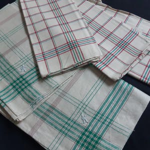 Pode incluir: Vários lenços brancos com um padrão xadrez em vermelho e verde. Alguns lenços têm uma borda verde e um pequeno desenho bordado. Os lenços estão dobrados e empilhados sobre uma superfície escura.