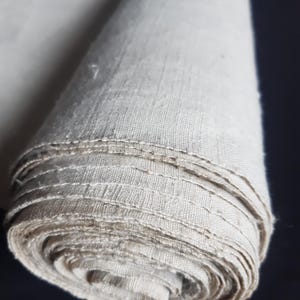 Pode incluir: Um rolo de tecido bege claro enrolado. O tecido parece ser linho ou um material tecido semelhante, com textura visível e um brilho sutil. As bordas do rolo mostram as camadas do tecido.