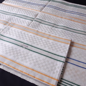 May include: A collection of white linen napkins featuring a checkered design and stripes in blue, green, and orange. The napkins are folded, with a dark background.