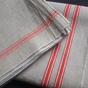 Pode incluir: Duas toalhas de linho cinzentas dobradas com riscas vermelhas e brancas. As toalhas estão empilhadas, mostrando as riscas vermelhas nas bordas. O tecido tem um aspeto texturizado.