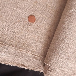 Pode incluir: Um close-up de um pedaço de tecido de linho natural, dobrado ao meio. O tecido é de cor bege claro e tem uma superfície áspera e texturizada. Um centavo de cobre está sobre o tecido.