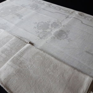 May include: A set of white linen napkins with a floral pattern. The napkins are folded and stacked on top of a larger white linen tablecloth with a similar floral pattern.