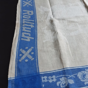 Pode incluir: Toalha de linho creme com uma borda azul e a palavra "Rolltuch" em azul. A borda apresenta padrões florais e geométricos. A toalha tem um design em relevo sutil.