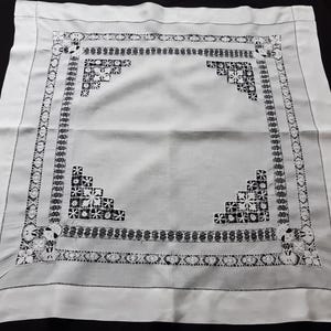 Op de afbeelding: Witte vierkante tafelkleed met ingewikkelde zwarte borduursels. Het patroon omvat bloemen en geometrische ontwerpen, een herhalende rand en bloemmotieven in de hoeken. De stof lijkt glad, mogelijk linnen.