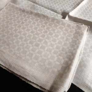May include: A stack of folded white linen napkins with a delicate chequered design. The napkins appear soft and textured, indicating a high-quality fabric. The square pattern adds a touch of elegance. The napkins are neatly arranged on a dark surface.