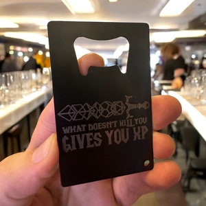 May include: Black metal bottle opener with a  'What doesn't kill you gives you XP' inscription and a sword and dice design.