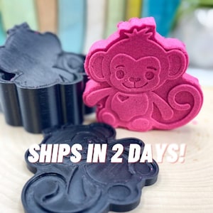May include: A pink monkey-shaped item and two black monkey-shaped molds. The monkey has a smiling face and is sitting. The molds are in the shape of the monkey. The background is blurred.