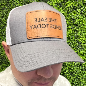 May include: Gray and white trucker hat with a leather patch. The patch features the text "THE SALE ENDS TODAY" in a brown leather rectangle. The hat has a gray brim and a white mesh back.