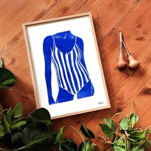 Peut inclure: Une impression encadrée représentant un maillot de bain à rayures bleues et blanches. L'œuvre est une linogravure de style minimaliste. Le cadre est en bois clair. L'image est sur une surface en bois.