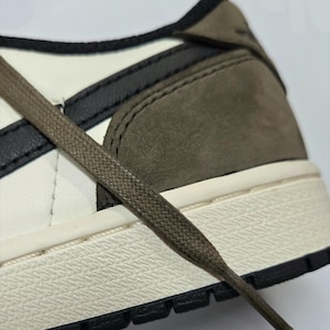 Può includere: Primo piano di una scarpa in pelle bianca e marrone con uno swoosh nero e lacci marroni. La scarpa ha un'intersuola bianca e una suola esterna nera.