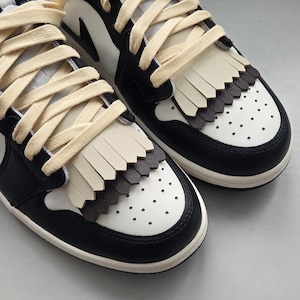Puede incluir: Un par de zapatillas deportivas negras y blancas con cordones color crema y flecos. Los zapatos tienen un diseño clásico con un toque moderno, con una puntera perforada y una combinación de colores contrastantes.
