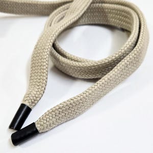 May include: Close-up of a beige braided drawstring with black plastic aglets. The drawstring is looped and lies on a white surface. The cord is thick and appears to be made of a durable material.