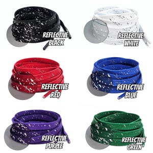 Op de afbeelding: Zes paar veters met reflecterende witte stippen op een gekleurde achtergrond. De kleuren zijn zwart, wit, rood, blauw, paars en groen. Elk paar heeft een label met de kleur en het woord "Reflectief" erop.