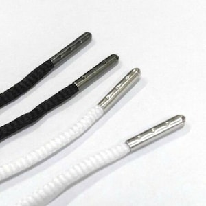 May include: Close-up of black and white shoelaces with silver metal aglets. The laces are made of a woven material and are displayed against a white background. The aglets have three small holes.