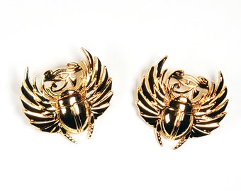 May include: A pair of gold-tone earrings featuring a scarab beetle design with wings. The beetles are detailed with intricate carvings and have a shiny finish.