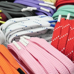 May include: A selection of shoelaces in various colours and patterns, including white with blue stitching, red with white stitching, pink, orange, and green. The shoelaces are arranged in a pile and some have plastic aglets.