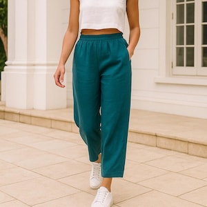 以下が含まれることがあります： エメラルドグリーンのリネンパンツ、ウエストはゴム仕様。白いクロップド丈トップスと白いスニーカーを合わせています。モデルはタイルの上を歩いています。