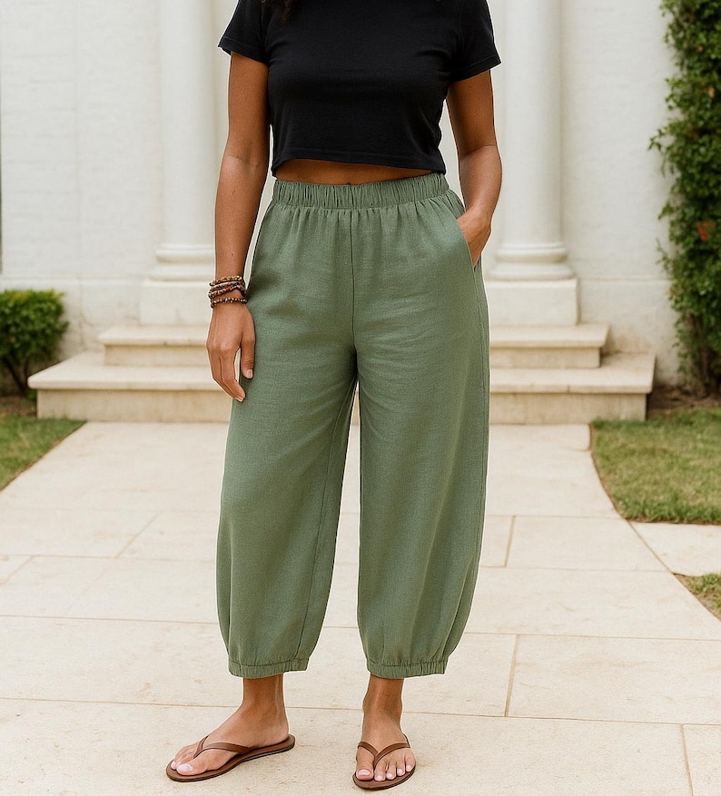 Op de afbeelding: Olijfgroene cropped broek met een elastische tailleband en geplooide elastische boorden bij de enkels. Deze wijde broek heeft zijzakken en wordt gecombineerd met een effen zwart T-shirt met korte mouwen en bruine teenslippers.