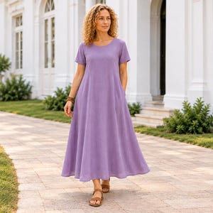 May include: A solid lavender midi dress featuring short sleeves, a round neckline, and a relaxed, flowing skirt silhouette. The garment is constructed with a central vertical seam detail running down the front bodice and includes side pockets.