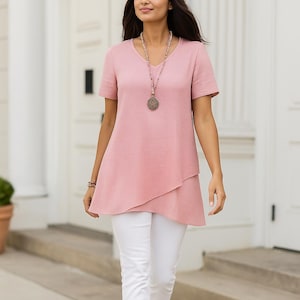 May include: A woman wearing a pink V-neck tunic top with short sleeves and white pants. The top has a layered asymmetrical hemline. A long beaded necklace and a bracelet are visible. The background features a white building with a door.