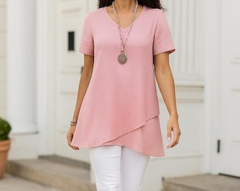 Op de afbeelding: Een vrouw draagt een roze V-hals tuniek met korte mouwen en een witte broek. De top heeft een gelaagde, asymmetrische zoom. Een lange kralenketting en een armband zijn zichtbaar. De achtergrond toont een wit gebouw met een deur.