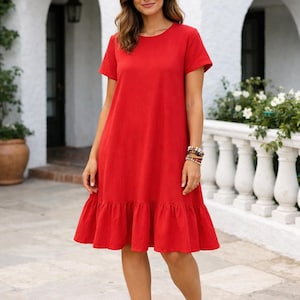 May include: A bright red, short-sleeved dress with a round neckline and a ruffled hem. The dress is knee-length and is paired with brown sandals. The background features a white building with arched doorways and a white balustrade.