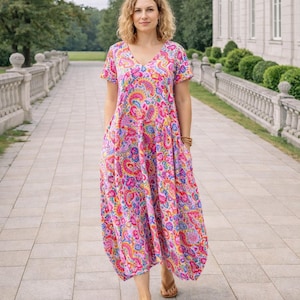 Op de afbeelding: Een vrouw draagt een roze paisley-jurk met een V-hals en korte mouwen. De jurk heeft een vloeiende, midi-rok en zakken. Ze draagt bruine sandalen en loopt op een geplaveide weg.