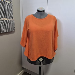 May include: A bright orange short-sleeved top with a round neckline and a textured, crinkled fabric finish. The garment features a loose, flowing silhouette with wide sleeves that drape naturally over the shoulders and sides of the torso.