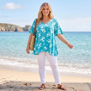Puede incluir: Una mujer en la playa lleva una blusa turquesa con estampado floral blanco y pantalones blancos. Lleva un bolso de mano tejido y sandalias marrones. El mar y el cielo azul se ven al fondo.