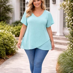 May include: A light blue V-neck top with short, flared sleeves and a layered design. The top is paired with blue jeans. The woman is smiling and walking on a stone path in front of a white building with arched windows.