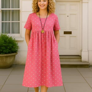 May include: A pink midi dress with a white floral pattern. The dress has short sleeves and pockets. A woman is wearing the dress with white sandals and a gold pendant necklace. The background includes a white door and window.