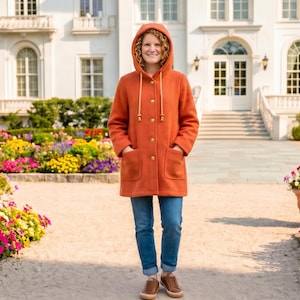 Puede incluir: Una persona con un abrigo naranja con capucha, bolsillos grandes y botones de madera, combinado con vaqueros azules y zapatos marrones. La persona está de pie frente a un edificio blanco con un jardín lleno de flores coloridas.
