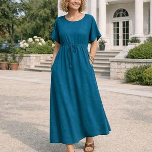 Puede incluir: Un vestido largo de color turquesa con mangas cortas y cintura con cordón. El vestido tiene bolsillos y se combina con sandalias marrones. La mujer está de pie frente a un gran edificio blanco.