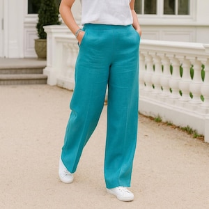 May include: High-waisted wide-leg trousers in a vibrant teal colour, featuring a clean waistband, side pockets, and a straight-cut silhouette. The trousers are paired with a simple white short-sleeved top and white lace-up trainers for a casual look.