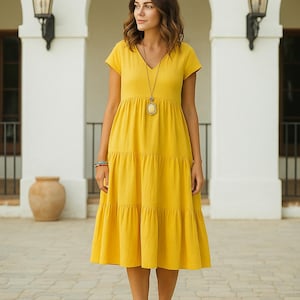 May include: A yellow tiered dress with a V-neck and short sleeves. The dress is paired with brown sandals and a long necklace. The background features a white building with arched doorways and decorative lamps.