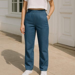 May include: Blue denim pants with a high waist and a straight leg. The pants have a classic five-pocket design and are paired with white sneakers. The model is wearing a white crop top and has a bracelet on their wrist.