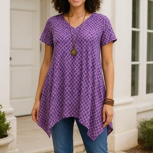 May include: A woman wearing a purple V-neck tunic with a geometric pattern, paired with blue jeans. She is also wearing a necklace and a bracelet. The tunic has short sleeves and an asymmetrical hemline.