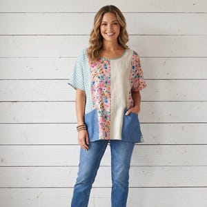 May include: A short-sleeved top featuring a patchwork design with a light blue gingham pattern, a floral print on a pink background, and a solid beige panel. The garment includes two blue patch pockets on the front and is paired with blue jeans.
