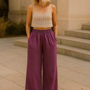 Puede incluir: Una mujer con un top corto de crochet beige con tirantes finos y pantalones morados de pernera ancha y talle alto. El conjunto se combina con sandalias. El fondo presenta un edificio y escaleras.