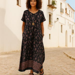 Puede incluir: Vestido negro de manga corta con cuello en V y estampado de cachemira en tonos marrones. El vestido tiene bolsillos y un borde decorativo en el dobladillo. La persona lleva un collar largo y sandalias. El vestido es adecuado para ropa informal.