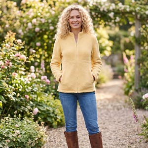 Könnte beinhalten: Eine Person trägt eine hellgelbe Fleecejacke mit Reißverschluss, blaue Jeans und braune Stiefel. Sie steht auf einem Kiesweg in einem Garten mit blühenden Blumen und Grün.