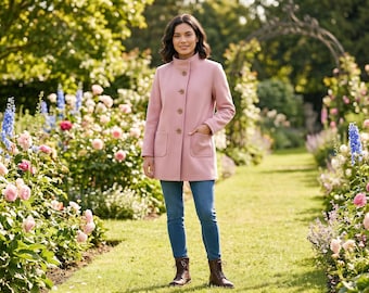 Op de afbeelding: Een vrouw draagt een lichtroze jas met een hoge kraag en knopen, gecombineerd met een blauwe jeans en bruine laarzen. De jas heeft twee voorzakken. De achtergrond toont een tuin met diverse bloemen en groen.