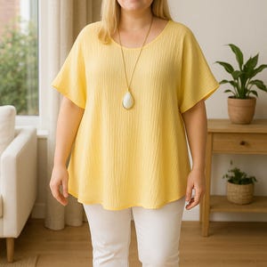 May include: A woman in a yellow short-sleeved top and white trousers. The top has a scoop neckline and a relaxed fit. A long gold necklace with a white pendant is worn. The outfit is casual and comfortable, suitable for everyday wear.