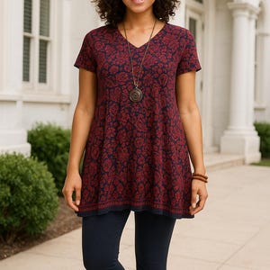May include: A woman wearing a V-neck tunic and dark blue leggings. The tunic is navy blue with a red floral pattern. A long necklace and a bracelet are visible. The background is a white building with columns.