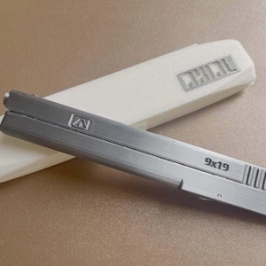 Könnte beinhalten: Ein grauer 9x19 Pistolenverschluss mit einem weißen Gehäuse. Der Verschluss trägt den Text "9x19" und ein Logo. Das weiße Gehäuse trägt den Text "DHILAL".