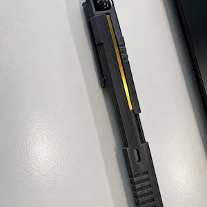 May include: A black and gold pen-shaped object with a small black sphere at the top. The pen has a gold stripe down the centre and a clip on the side. The bottom of the pen has a series of horizontal lines.